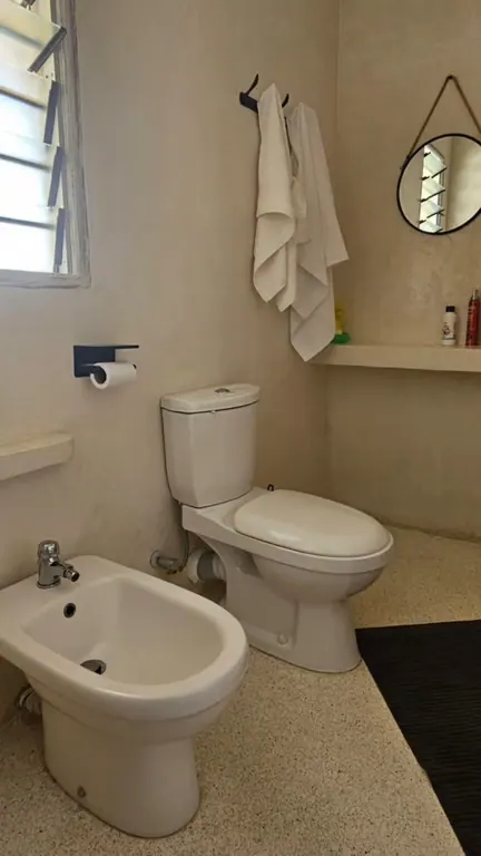 Compact bathroom with toilet, bidet, round mirror, shelf and louvred window