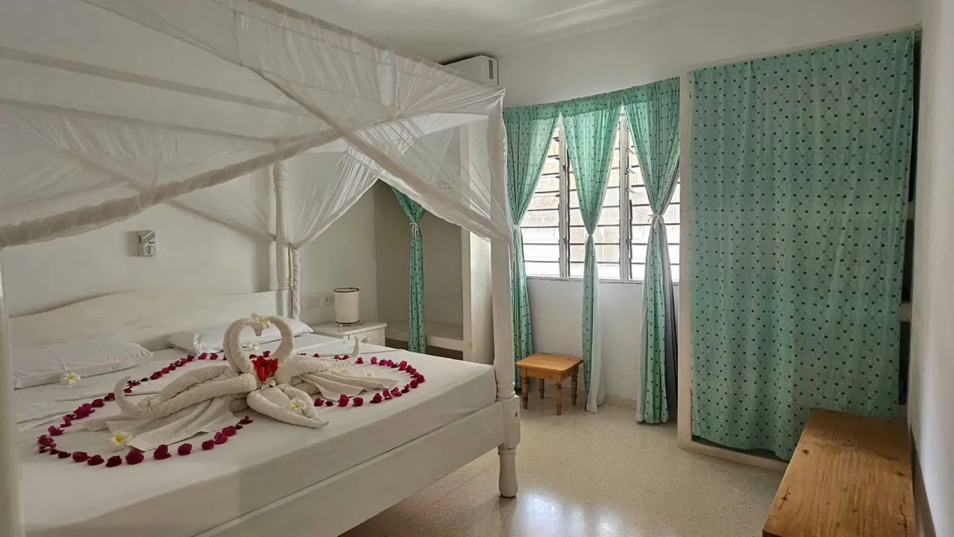 White four-poster bed beneath a mosquito net beside aqua curtains and a window