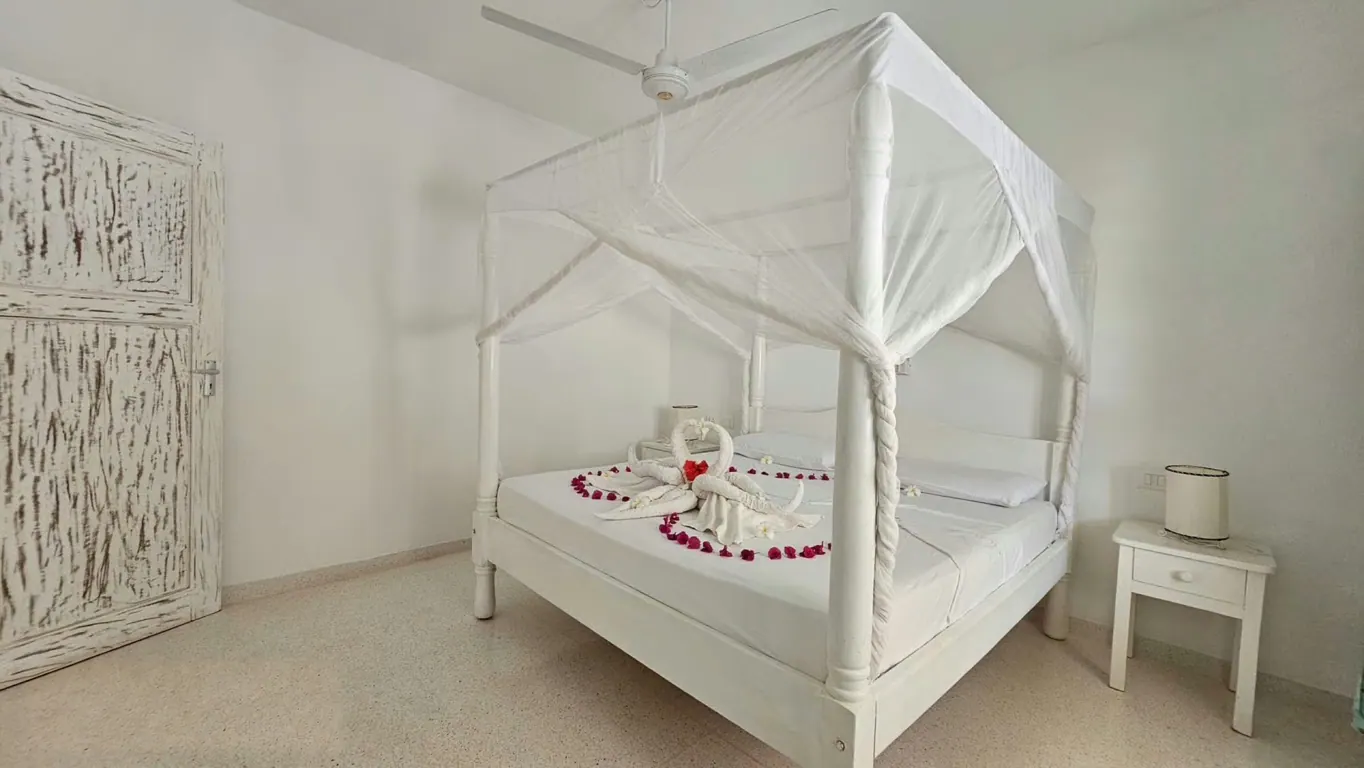 White four-poster bed beneath a mosquito net with a towel arrangement and red petals
