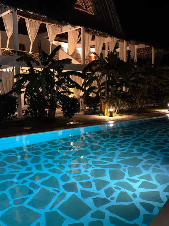 Illuminated white thatched building behind the blue-lit shared pool at night