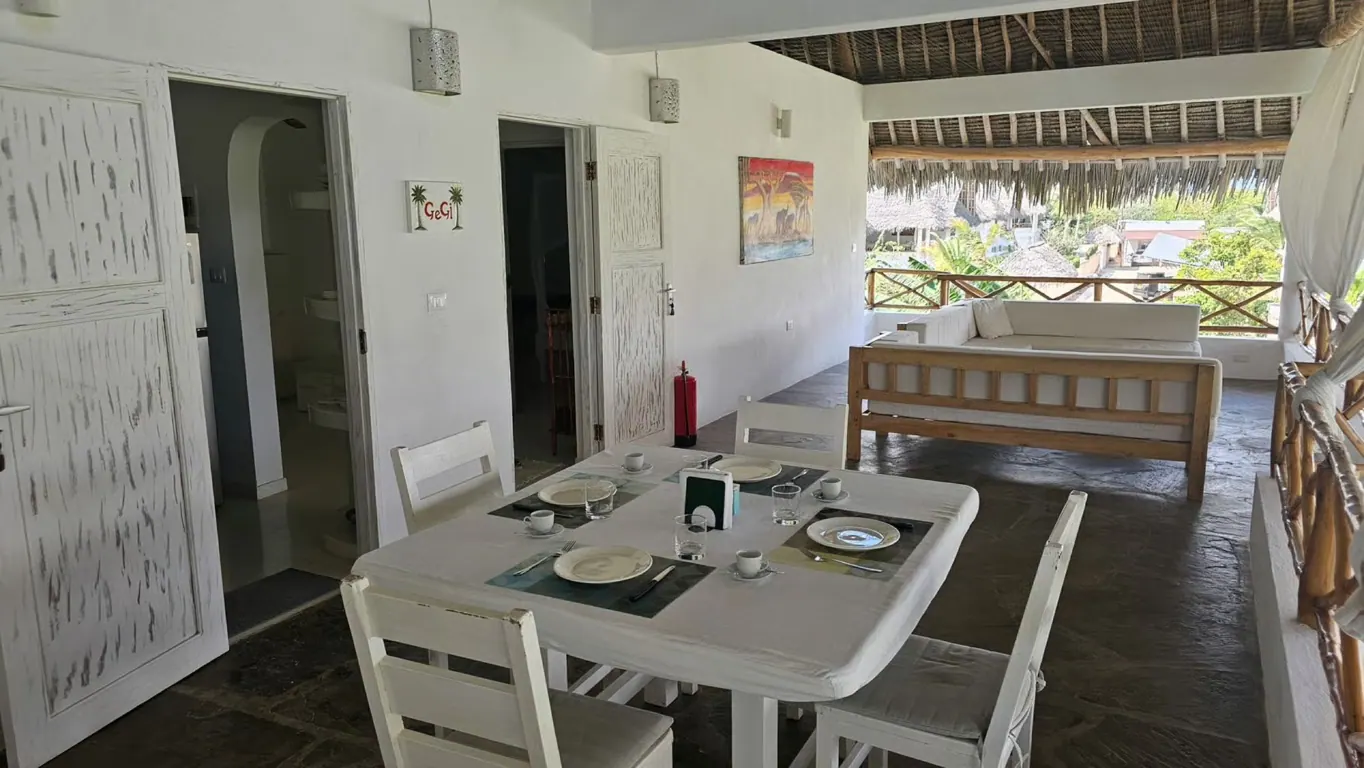 Dining table set for four on a covered terrace, with white doors and a lounge behind