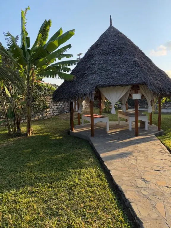 Thatched open pavilion with two massage tables beside a stone path and banana plants