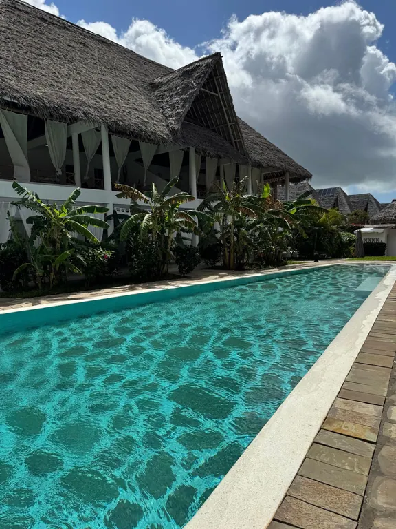 Long turquoise shared pool beside a white thatched building and banana plants