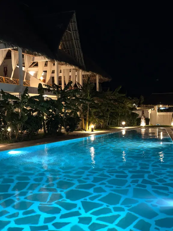 Long blue-lit shared pool beside an illuminated thatched building at night